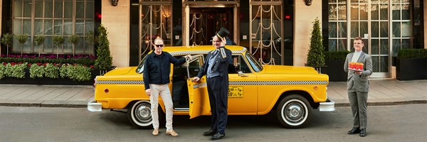 A yellow vintage taxi parked outside Claridge’s, with a guest, a doorman tipping his hat, and a server holding a tray of cocktails.