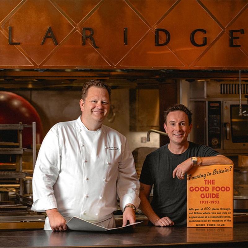 Two men are standing in a professional kitchen under a sign that reads