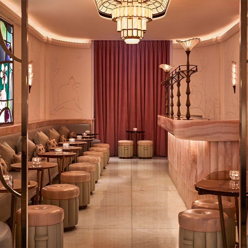 Elegant Art Deco-style lounge with pink onyx bar, soft lighting, round stools, and mauve velvet curtains.