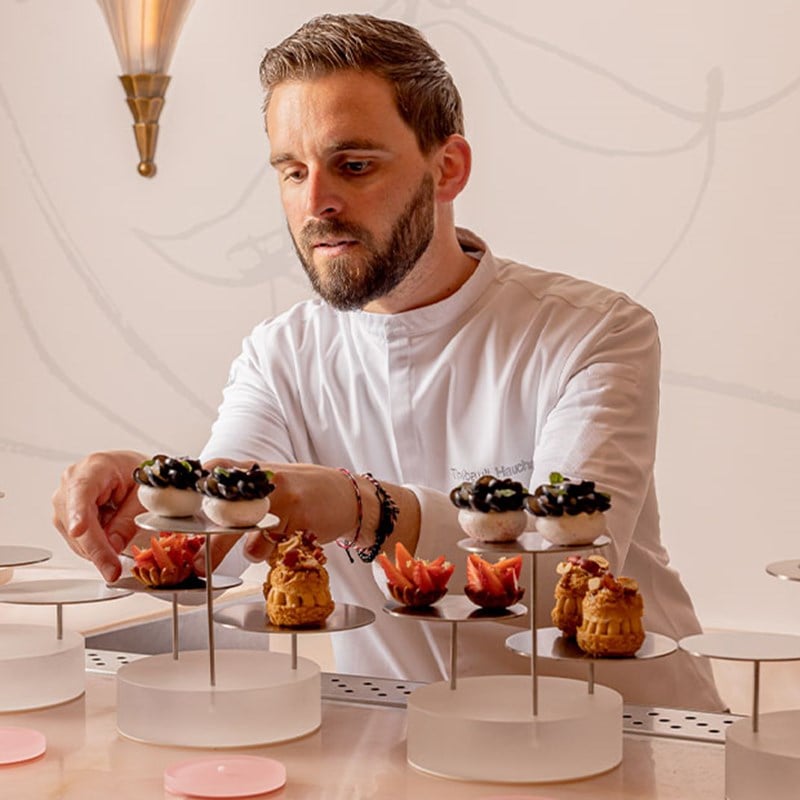 Pastry chef delicately arranges a selection of miniature desserts on modern glass stands in a softly lit setting.