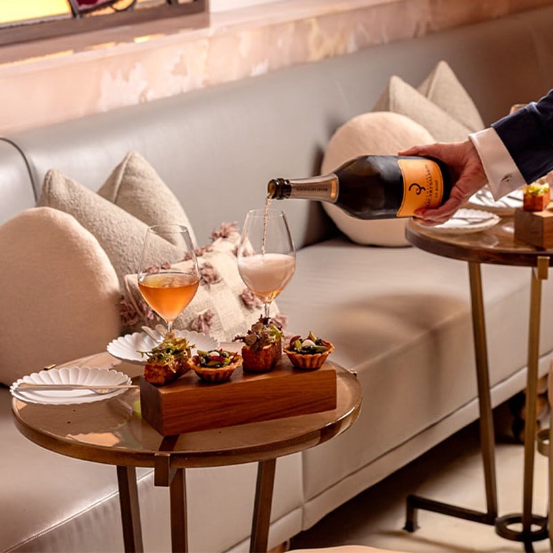 A server pours champagne beside a canapé platter on a small round table with plush sofa seating and neutral-toned cushions.