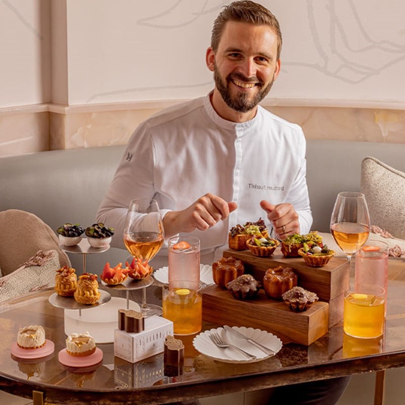 Smiling chef sits behind a luxurious afternoon tea display featuring savoury bites, desserts, and cocktails in an Art Deco setting.