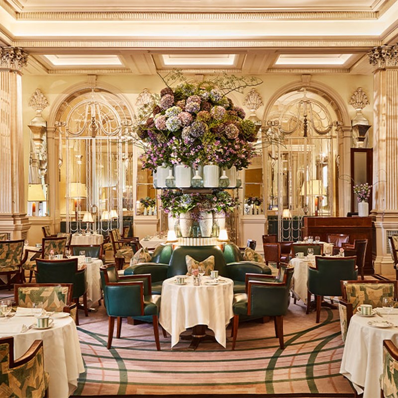 Opulent tea salon with green armchairs, mirrored walls, and a dramatic floral arrangement in the centre.