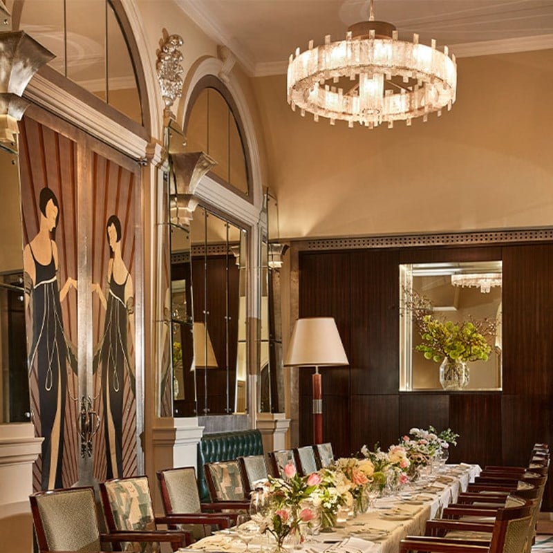 Elegant private dining room with a long flower-adorned table, mirrored walls, and Art Deco mural panels.