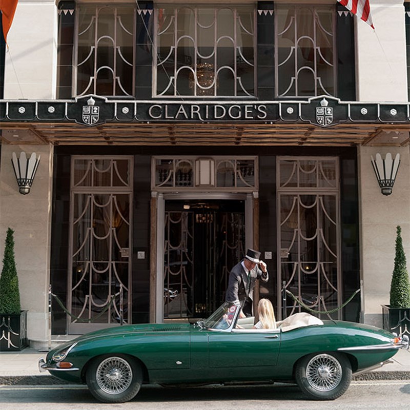 A doorman greets a guest in a vintage green convertible outside the ornate black-and-silver entrance of Claridge’s Hotel.