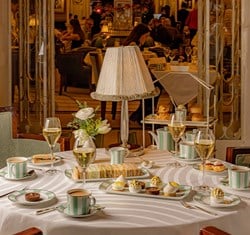 A round table elegantly set for afternoon tea with striped green china, tiered pastries, and champagne in a softly lit room.
