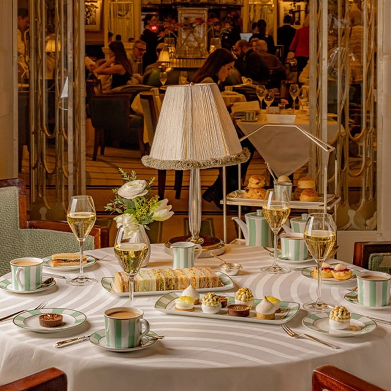 A round table elegantly set for afternoon tea with striped green china, tiered pastries, and champagne in a softly lit room.