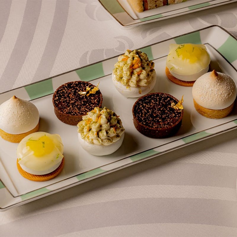 A rectangular plate with intricate mini desserts, including chocolate, citrus, and meringue pastries, on green-trimmed china.