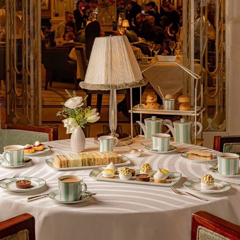 An elegant tea table with sandwiches, cakes, and tea service, surrounded by mirrored décor and softly lit diners.