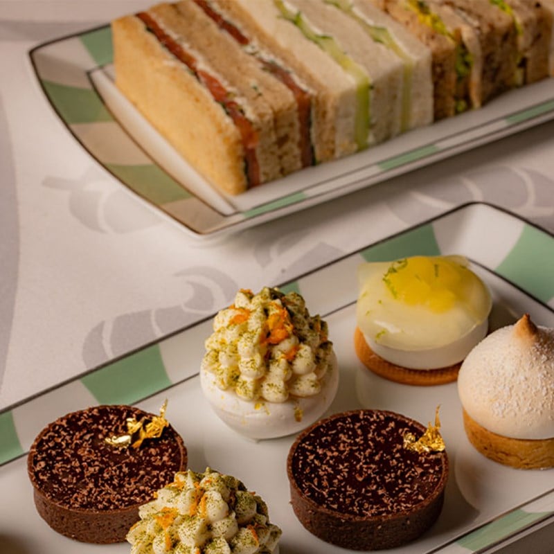 Neatly stacked finger sandwiches and ornate mini desserts presented on matching green-edged porcelain plates.
