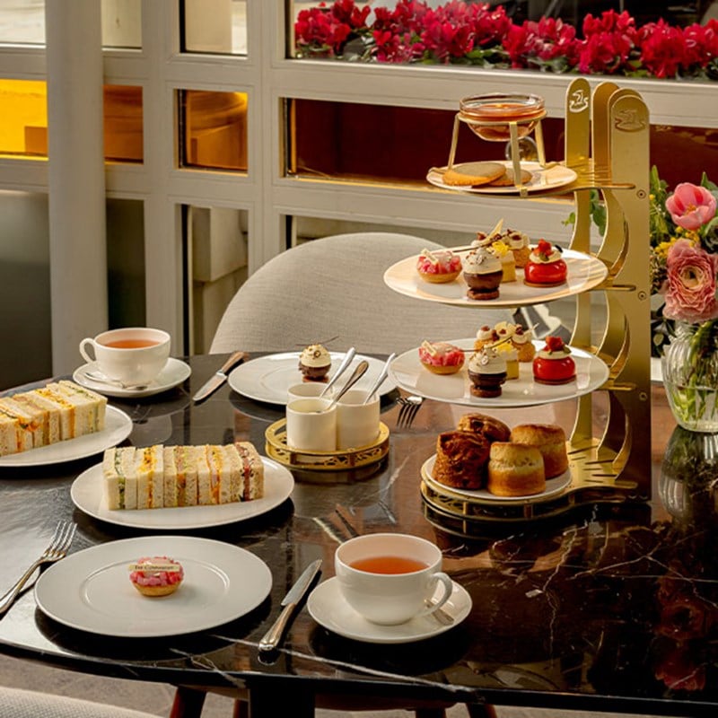 A marble table set for afternoon tea with a golden tiered stand filled with scones, cakes, and pastries next to fine china and fresh flowers.
