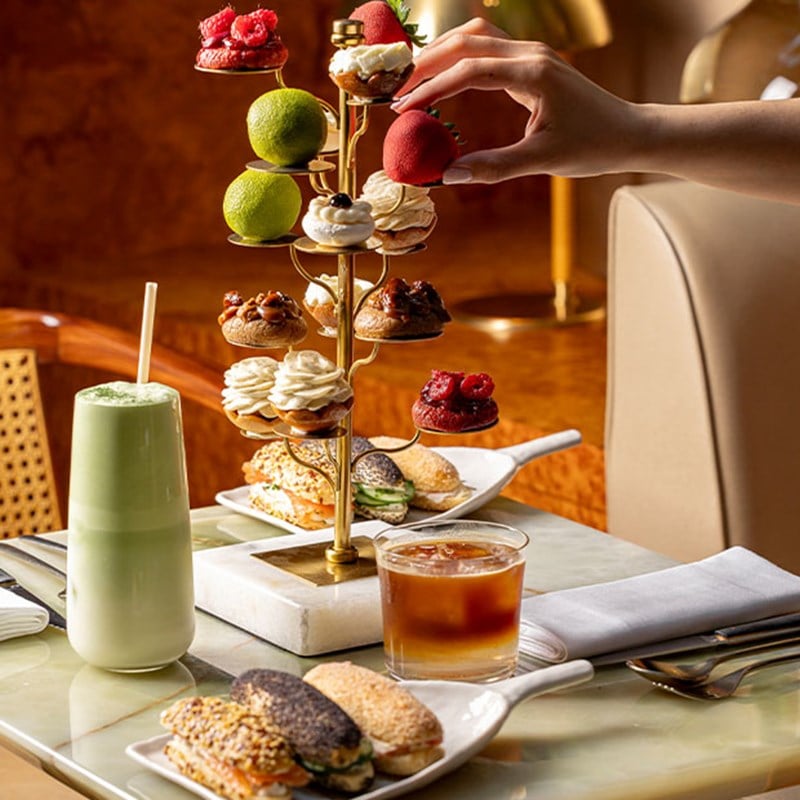 A unique tree-like stand holds colourful pastries above a selection of sandwiches and iced drinks on a modern café table.