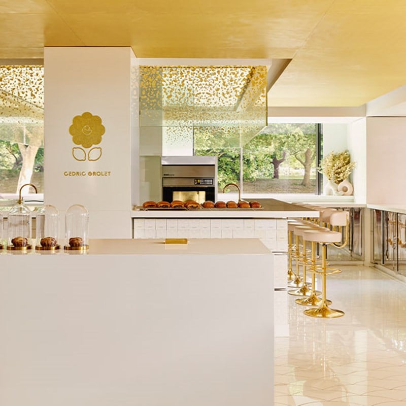Sleek, minimalist patisserie with golden accents, high stools, and pastries displayed under glass domes, with a view of greenery outside.