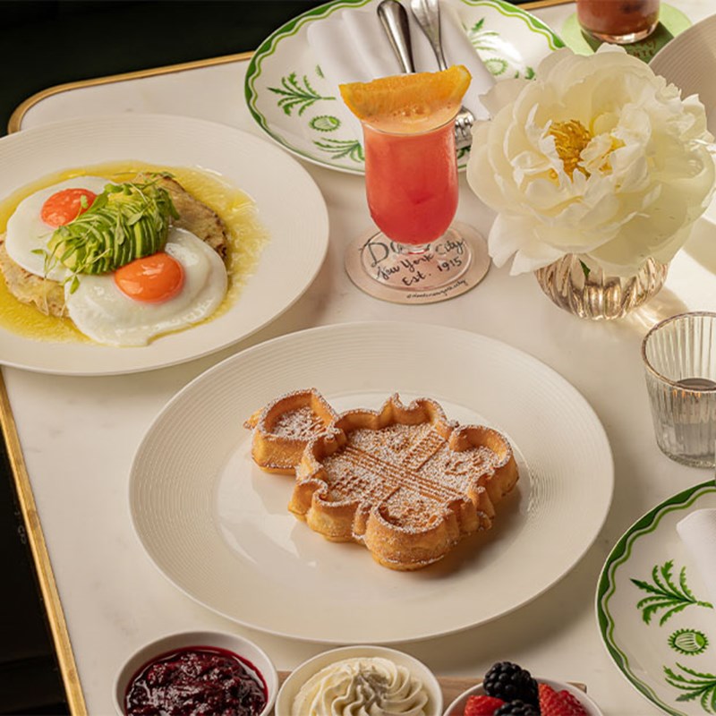 A white marble table set with a decorative waffle, sunny-side-up eggs with avocado, a pink cocktail, and a fresh white peony flower in a vase.