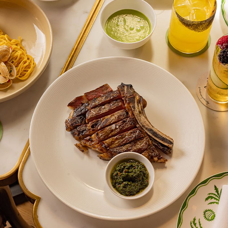A grilled bone-in steak sliced and served with green chimichurri sauce on a white plate, surrounded by pasta and green sauce.