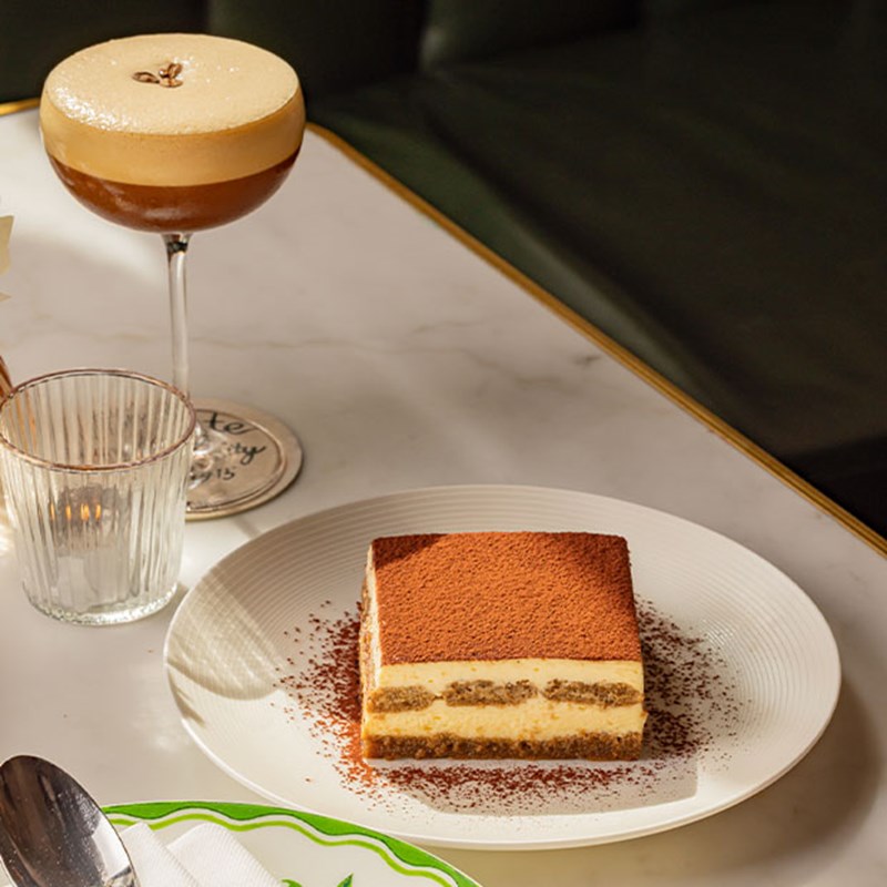 A square slice of tiramisu on a white plate, lightly dusted with cocoa, beside an espresso martini on a marble table.