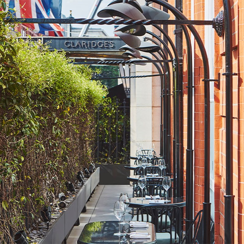 A narrow terrace lined with black tables and chairs under wrought iron arches, with Claridge’s signage visible in the background.