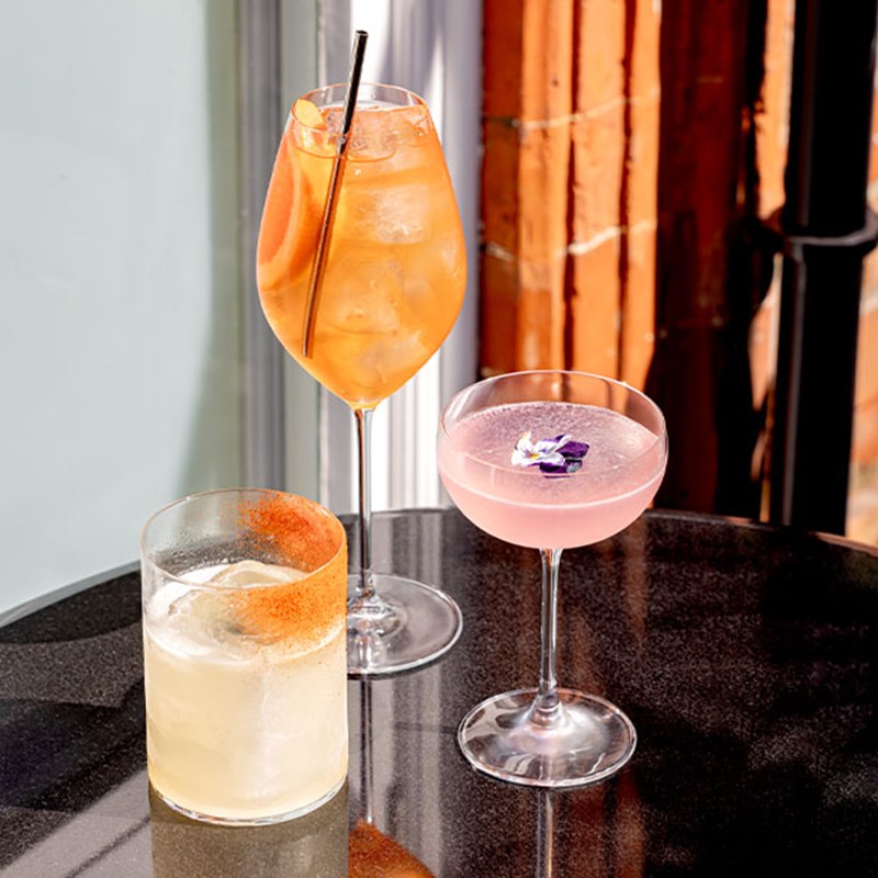 Three elegant cocktails on a round black table—one in a highball glass with ice, one in a coupe with a flower garnish, and one in a rocks glass with an orange rim.