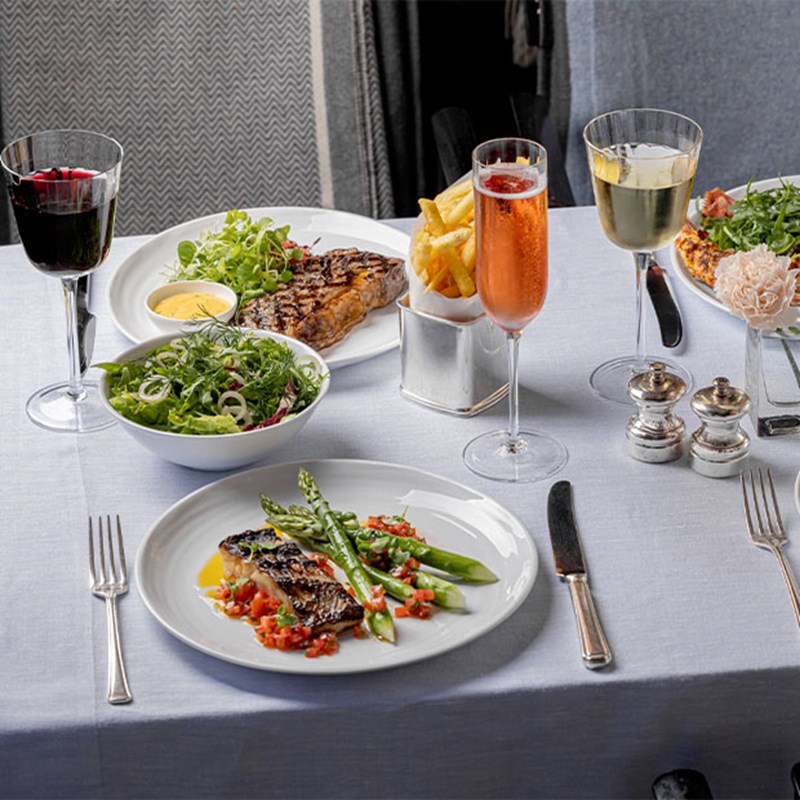 A white tablecloth dining setup with grilled fish, steak, fries, salads, and sparkling wine and cocktails, arranged for two.