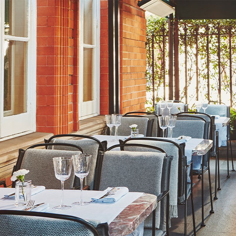 A shaded outdoor dining area with grey herringbone chairs and white-tablecloth tables next to a red brick building.