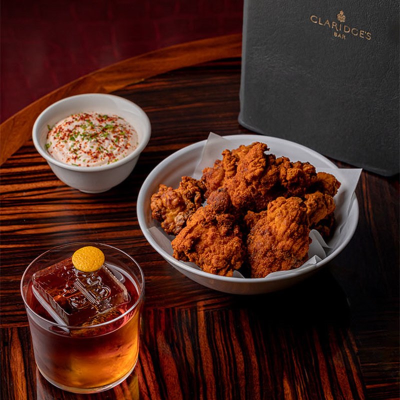A wooden table with a bowl of golden fried chicken, a small bowl of seasoned dip, and a dark cocktail with a round garnish.