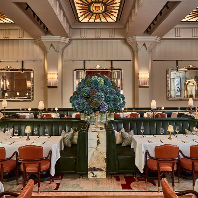 A grand dining room with art deco ceiling lights, green leather banquettes, white tablecloths, and a large central floral arrangement of hydrangeas.