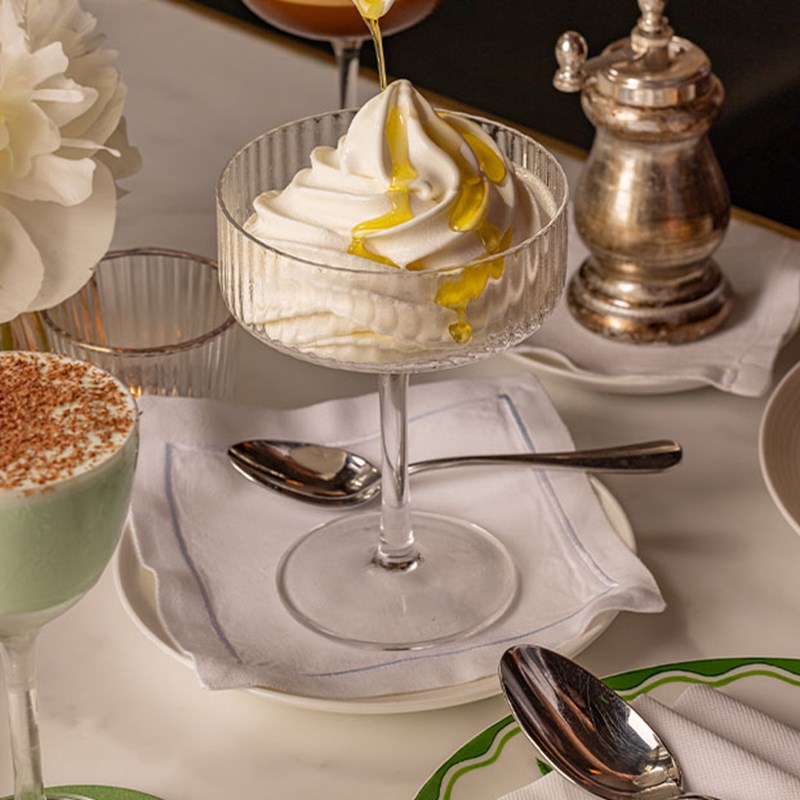 A swirl of vanilla soft-serve ice cream in a glass coupe, drizzled with olive oil, placed on a linen napkin with a silver spoon.