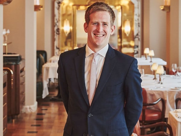 Smiling man in a navy blue suit and patterned tie stands confidently in an elegant, softly lit dining room with Art Deco décor and white tablecloths.