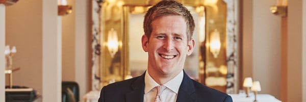 Portrait of a smiling man in a navy suit standing in an elegant, warmly lit dining room with Art Deco detailing.