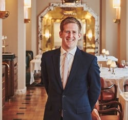 Portrait of a smiling man in a navy suit standing in an elegant, warmly lit dining room with Art Deco detailing.