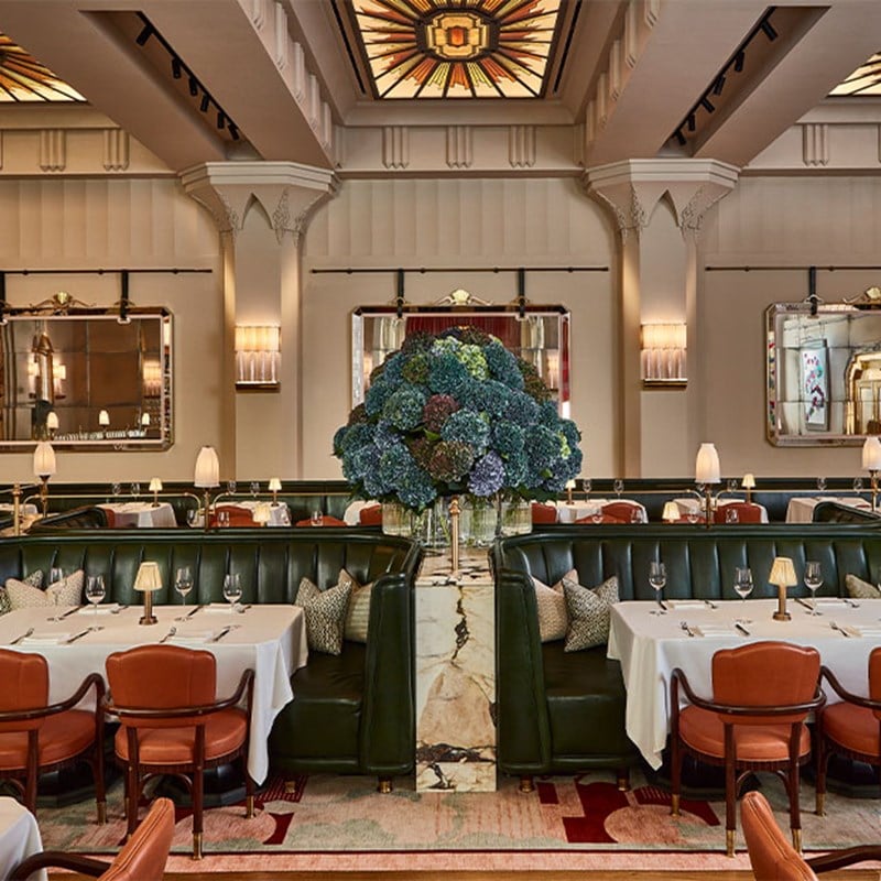 View of a grand dining room with green leather banquettes, orange chairs, and a central floral arrangement beneath a stained glass ceiling.