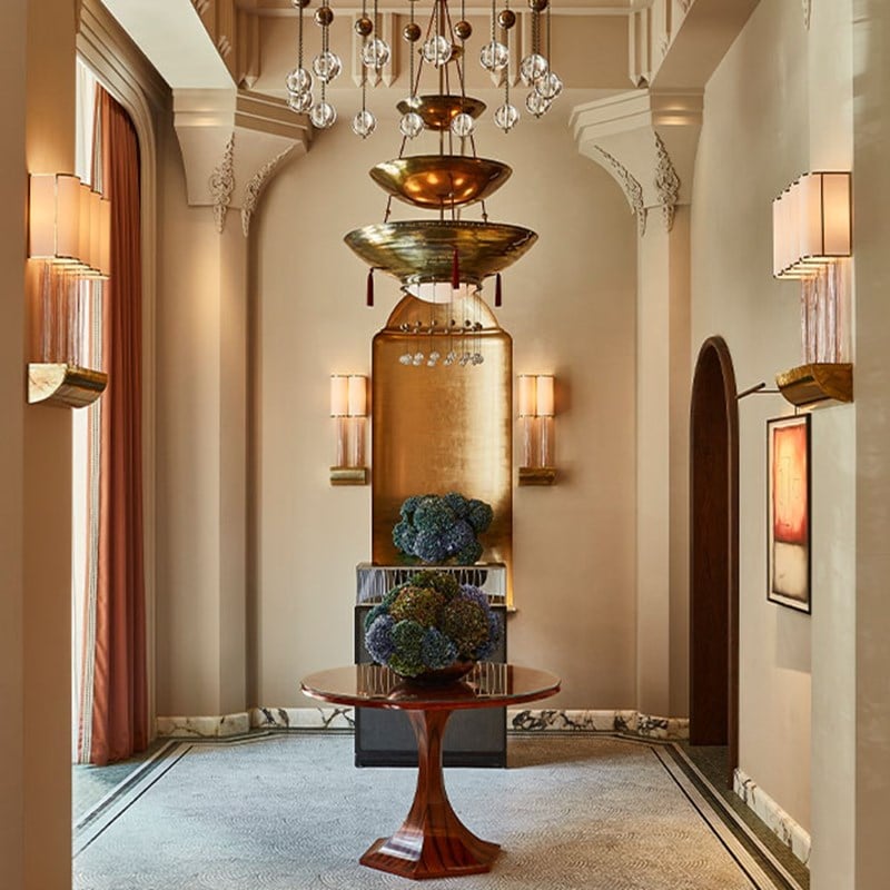 Ornate hallway featuring a sculptural golden chandelier, marble accents, and a round table with a floral centerpiece.
