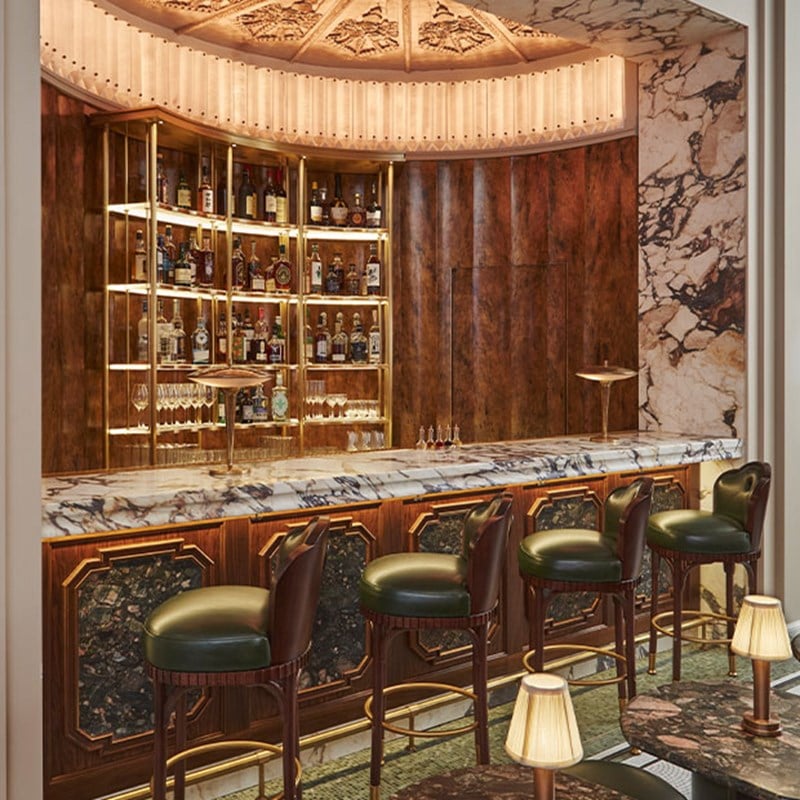 Elegant bar with a marble countertop, green leather stools, and a backlit display of spirits against a rich wood and stone backdrop.