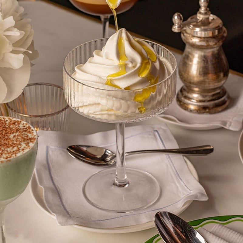 Close-up of a soft-serve ice cream dessert drizzled with olive oil, served in a glass coupe on a white napkin.