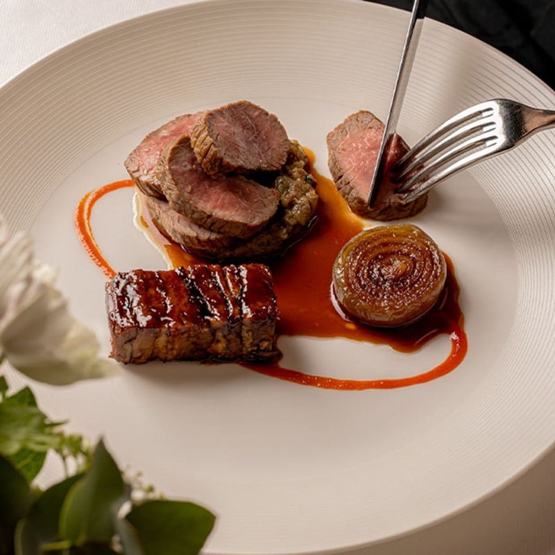 Plated gourmet dish featuring sliced lamb, glazed onion, and a square cut of braised meat with a rich sauce.