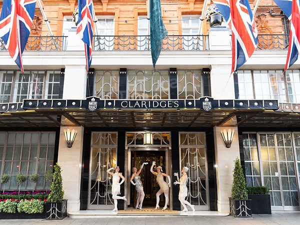 Four women dressed in 1920s-style flapper costumes pose playfully outside the entrance of Claridge’s hotel, which is decorated with Union Jack flags and black Art Deco detailing above the doors.