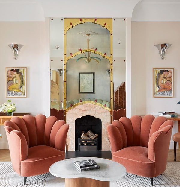 Art Deco-style sitting room at Claridge’s featuring two scallop-shaped, terracotta velvet armchairs positioned around a sculptural stone coffee table. The focal point is a decorative fireplace with a pink marble surround and an ornate, mirrored overmantel with gold and red detailing. Flanking the fireplace are abstract paintings and chrome wall sconces, with soft, neutral-toned walls and elegant furnishings completing the refined aesthetic.
