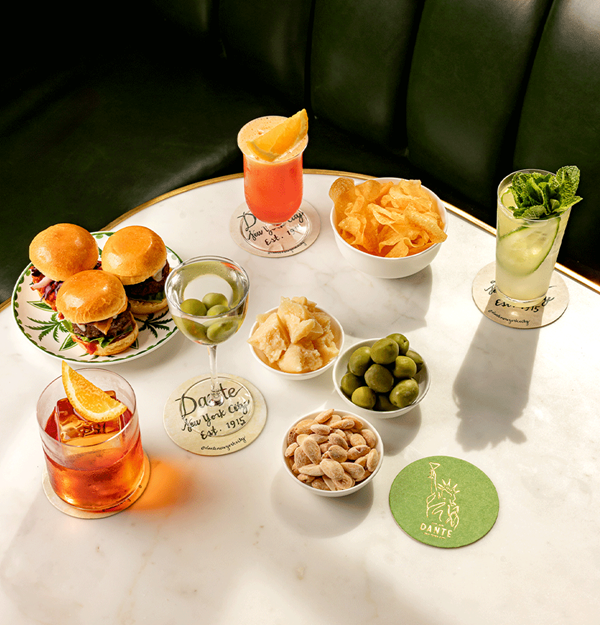A marble table set with three mini sliders on a patterned plate, bowls of green olives, potato chips, almonds, and cheese crisps, accompanied by four cocktails, each garnished with citrus or mint, at a booth with green leather seating.