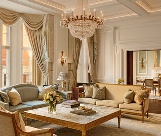 Elegant living room with crystal chandelier, tall draped windows, gold-trimmed furniture, and a marble coffee table, leading into a formal dining room beyond.