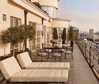Expansive private terrace with cream sun loungers, black metal dining set, potted olive trees and topiary, overlooking London’s skyline on a clear day.