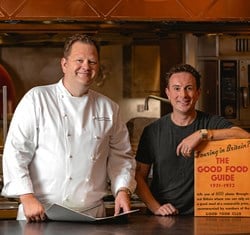 Two men are standing in a professional kitchen under a sign that reads