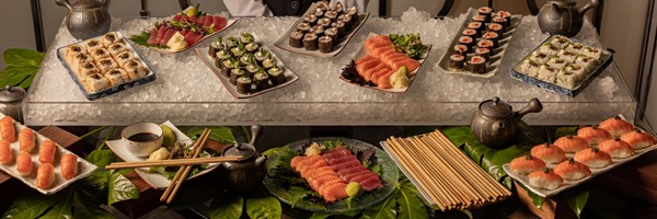Buffet spread of assorted sushi and sashimi arranged on ice, with soy sauce, wasabi, chopsticks, and teapots, attended by a server.