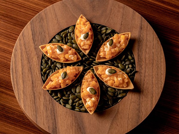 Wooden platter with five leaf-shaped pastry shells filled with finely diced orange vegetables, topped with cream and pumpkin seeds.