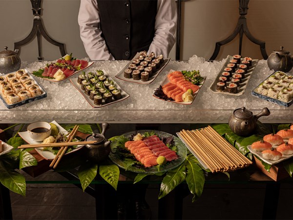 Buffet display of assorted sushi and sashimi on ice, with soy sauce, wasabi, chopsticks, and tea pots, attended by a server.