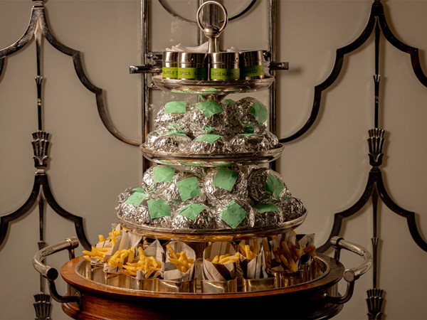 Three-tiered silver stand with foil-wrapped sandwiches, jars of seasoning, and paper cones of golden fries on a wooden serving trolley.