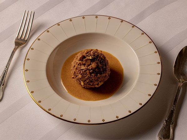 Dessert served in a deep-rimmed plate, featuring a round scoop coated in chocolate and nut crumble, resting in a pool of caramel-coloured sauce.