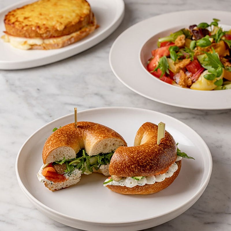 Smoked salmon and cream cheese bagel in foreground with croque monsieur and salad in background, all on a marble table