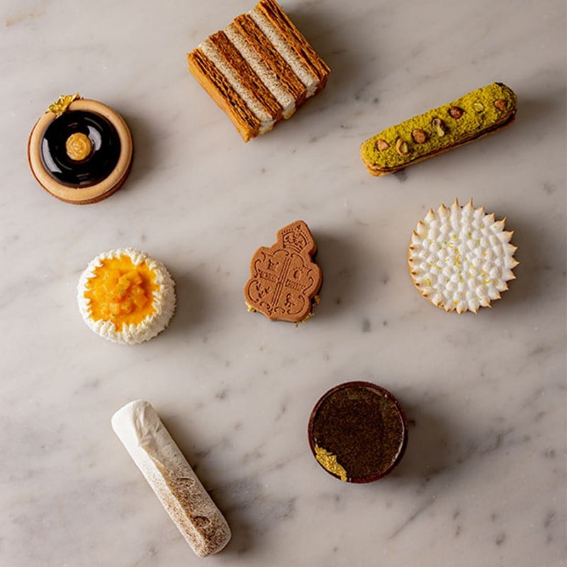 An elegant assortment of artisanal pastries, including éclairs, mille-feuille, citrus tarts, and a signature Claridge’s biscuit, beautifully displayed on a marble background.