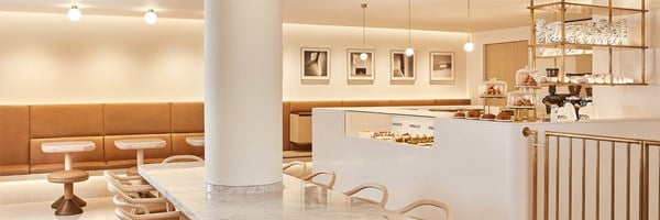Wide-angle view of the ArtSpace Café featuring a sleek white counter displaying pastries, caramel leather banquettes, marble-top tables, and a display of coffee cups and crockery on brass shelving.