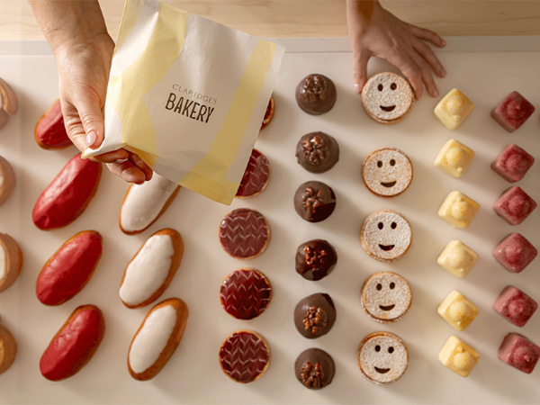 Hands select assorted pastries from a display, including iced cakes and chocolates, beside a Claridge’s Bakery paper bag.